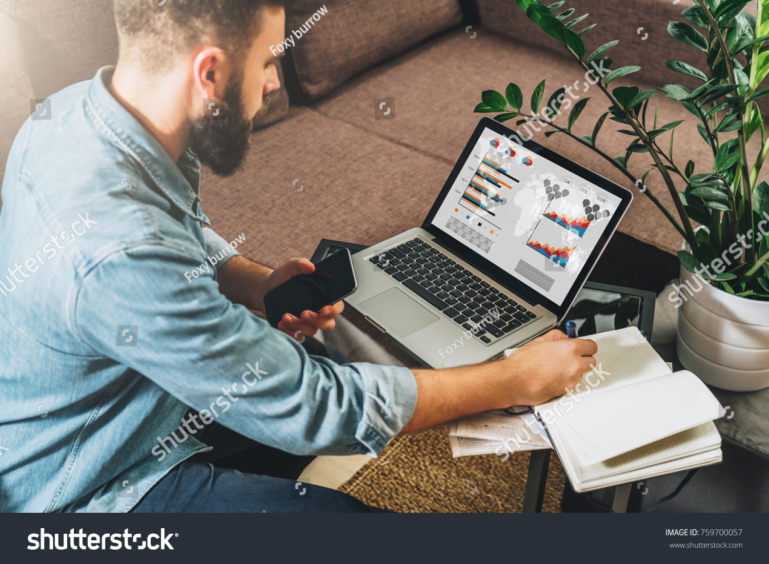 Man sits at table,working,holds smartphone,uses laptop with graphs, charts, diagrams on screen and makes notes in his notebook.Online marketing, education, e-learning, e-commerce. Startup, planning. #759700057