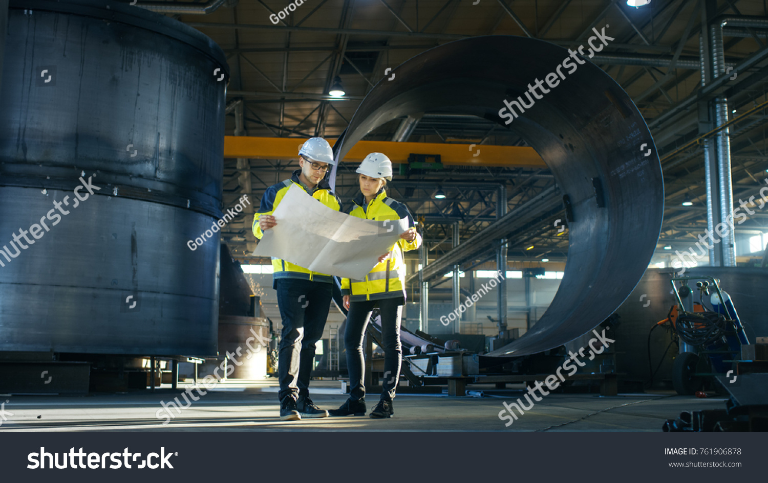 Male and Female Industrial Engineers Look at Project Blueprints While Standing Surround By Pipeline Parts in the Middle of Enormous Heavy Industry Manufacturing Factory #761906878