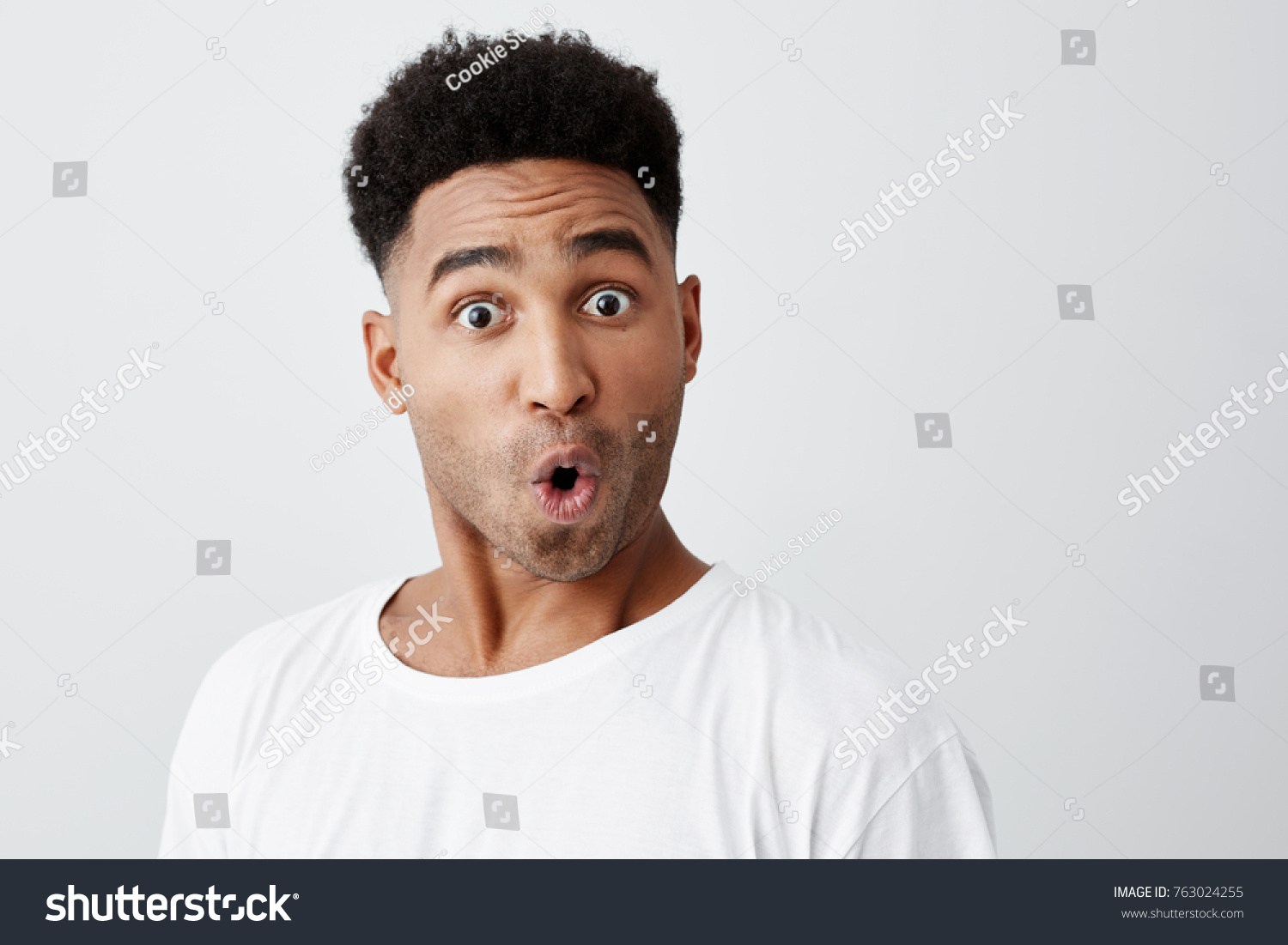 Close up of funny handsome young dark-skinned man with afro haircut in stylish white t-shirt looking in camera with raised eyebrows and surprised face expression. #763024255