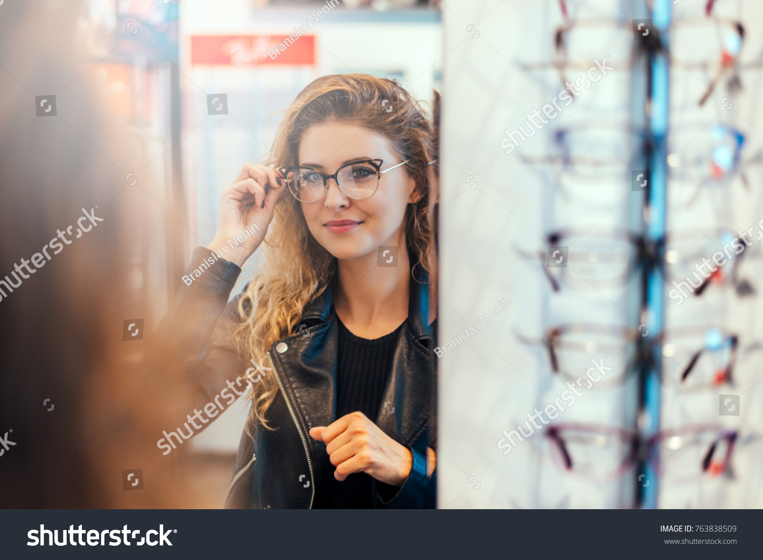 Smiling young woman trying on glasses on mirror in optician. #763838509