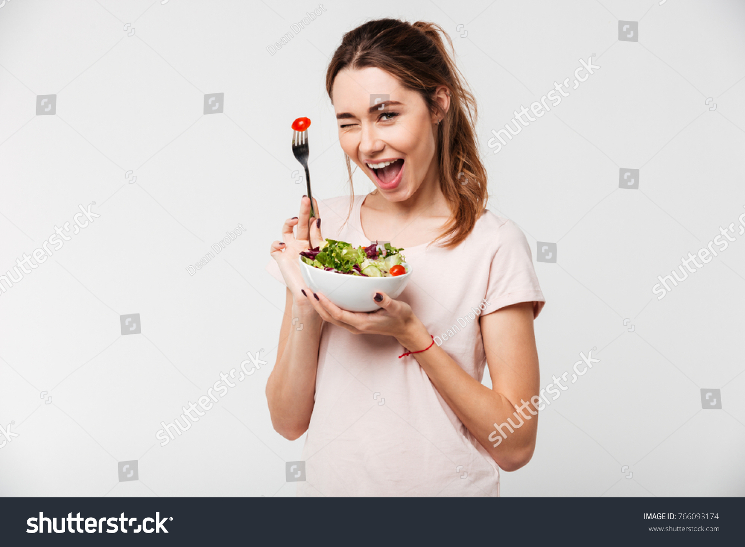 Portrait of a happy playful girl eating fresh salad from a bowl and winking isolated over white background