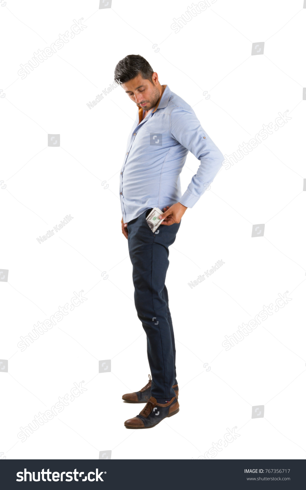 Black short hair young caucasian guy with blue shirt, blue casual pants and brown shoes counting usa dollar money isolated on white background.  #767356717