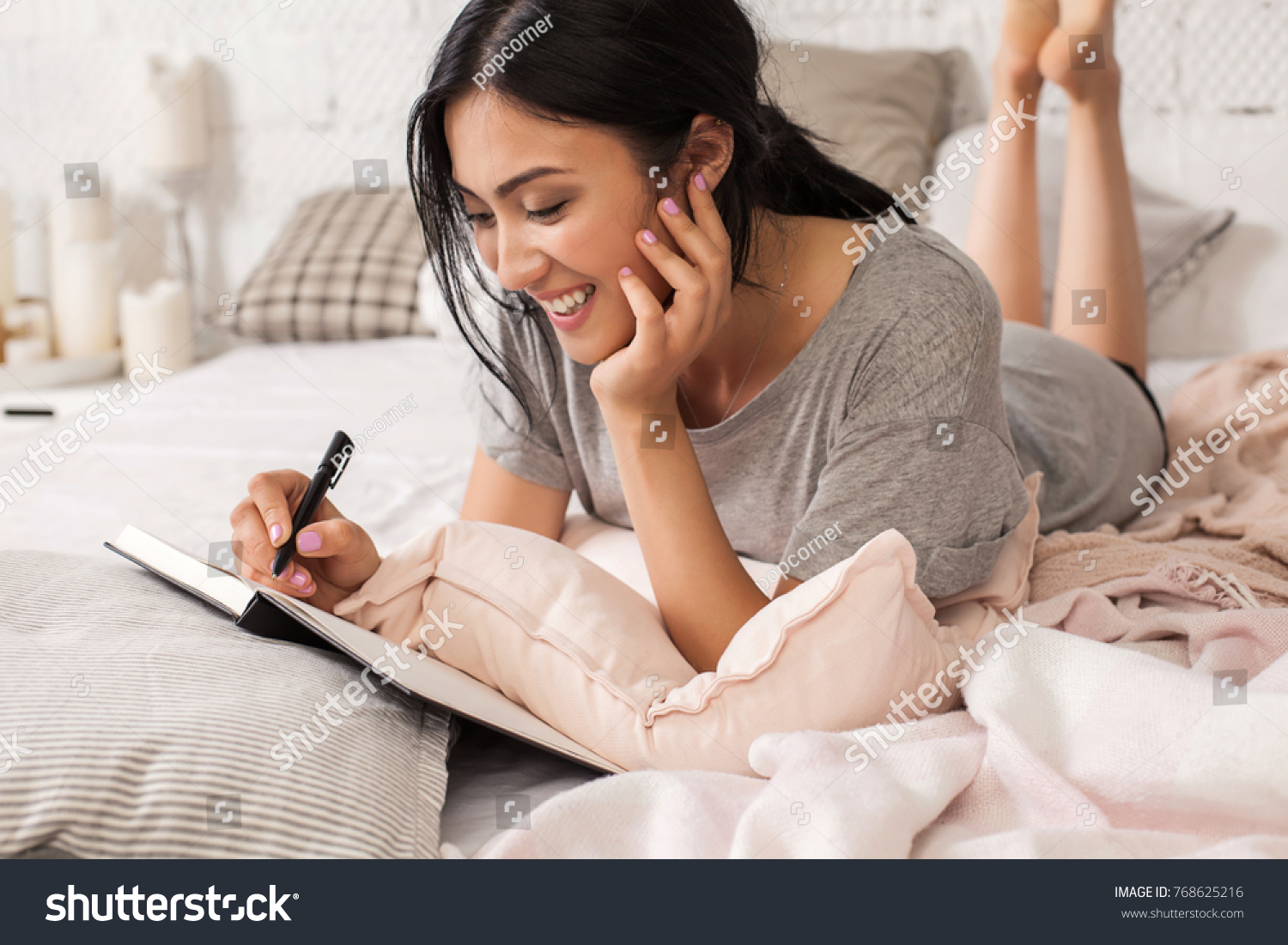 Beautiful young asian woman laying on bed and writing a diary. Smiling brunette lady with notebook and pen in her hands. Modern bright home interior on background. #768625216