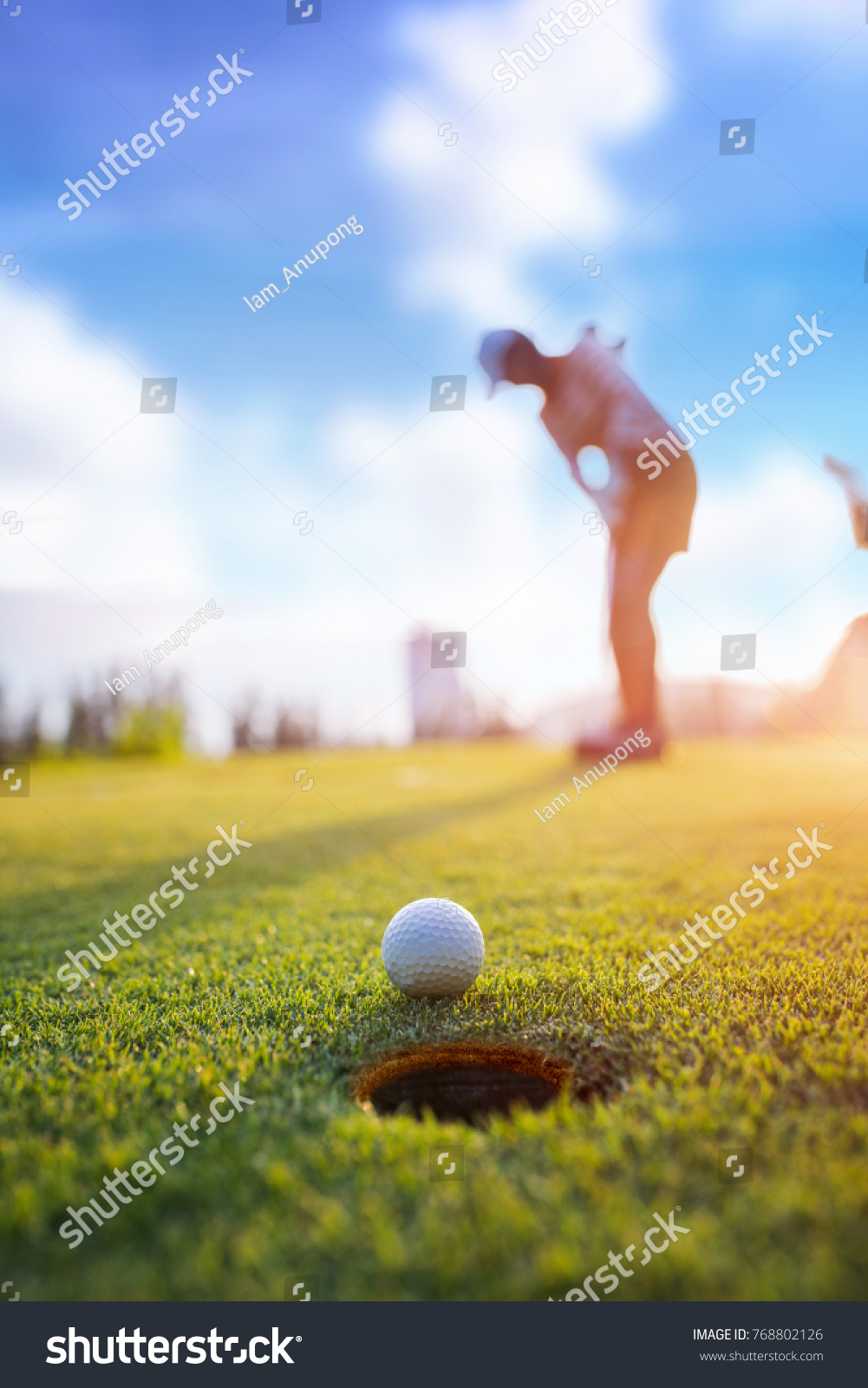 Golf ball putting by woman golf player in background, golf ball spining to the hold on the green of golf course with early light of sunset #768802126