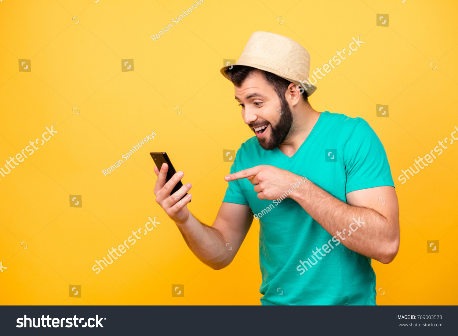 Ha-ha! So funny! Portrait of happy excited crazy man with stubble wearing hat and green tshirt, he is pointing on his smartphone and rejoicing #769003573