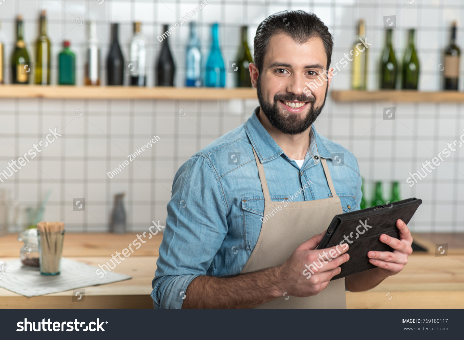 Convenient gadget. Positive emotional bearded waiter looking glad while standing with a modern convenient tablet in his hands and smiling #769180117