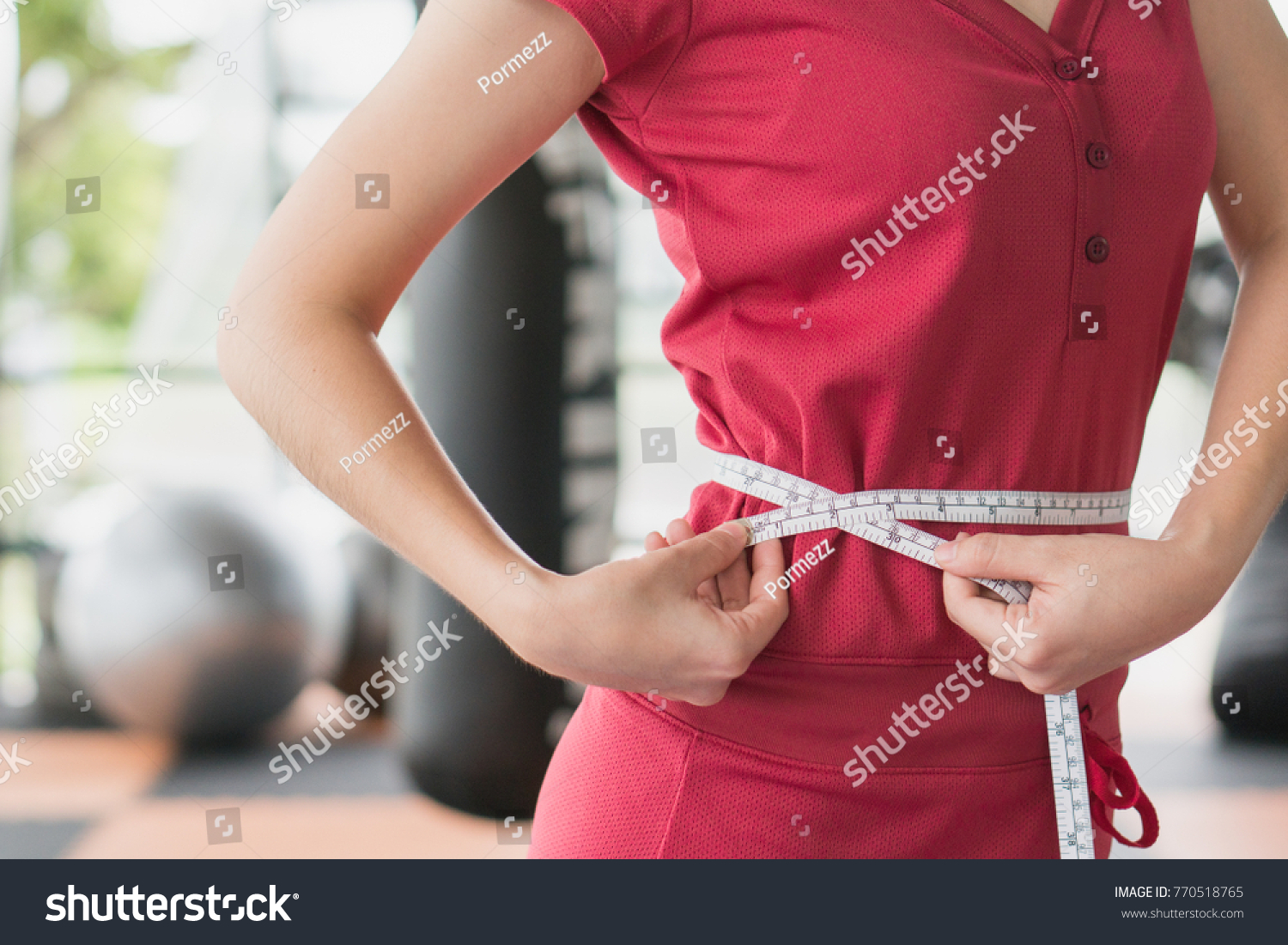 Young slim woman measuring her waist by measure tape after a diet with accessory in sporty gym as background. #770518765
