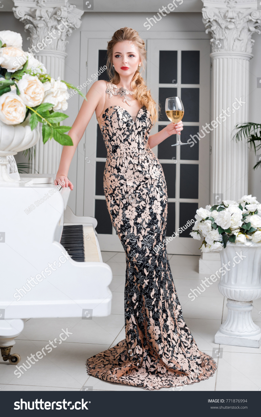 Elegant blonde lady with glass of wine in restaurant standing near white grand piano in a luxury classic interior. Beautiful sexy young woman with perfect body and pretty face make-up wearing evening #771876994