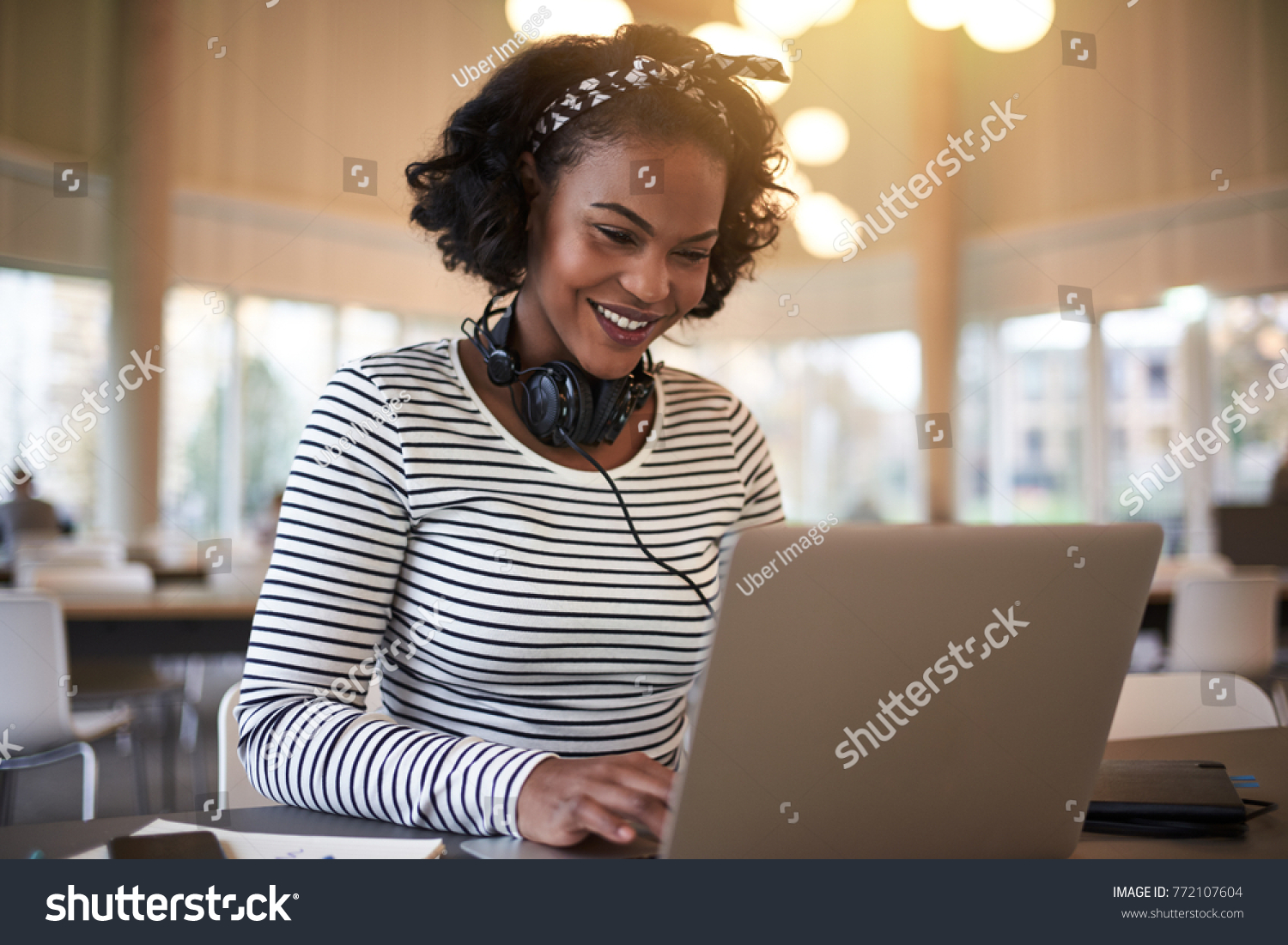 Smiling young African university student sitting on campus wearing headphones and working online with a laptop #772107604