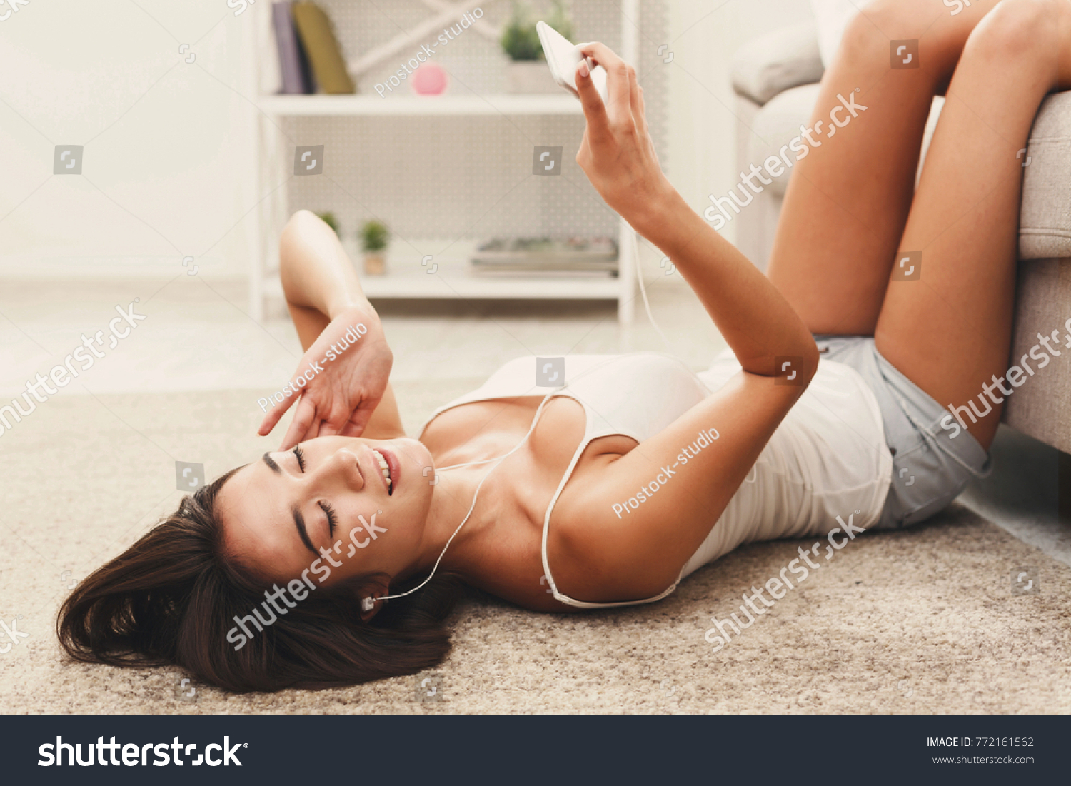 Young brunette woman enjoying the music in earphones and relaxing while lying at home on floor carpet, with her legs up on the sofa. Technology and enjoyment concept #772161562