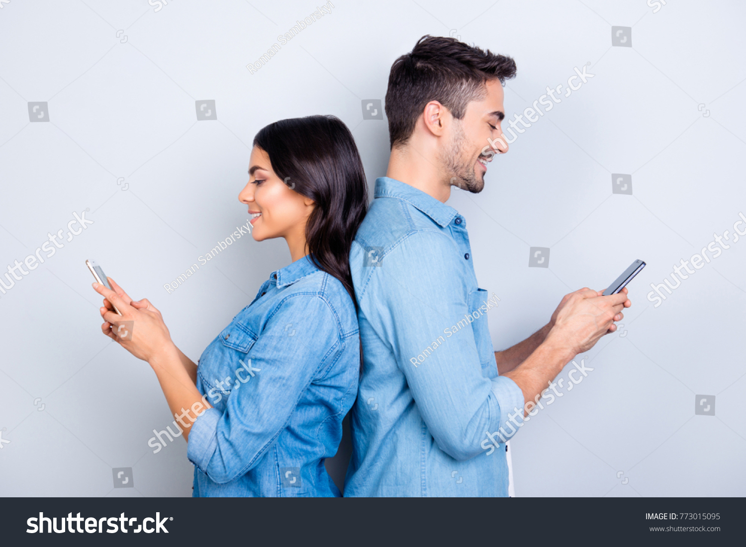 Beautiful, pretty, cute woman and handsome man in jeans shirts standing back to back and writing sms through 3g internet on smart phones over grey background #773015095