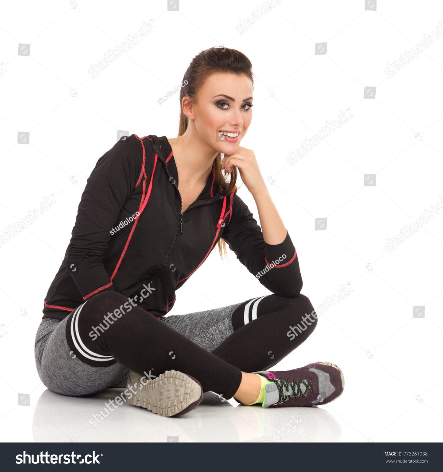 Beautiful young woman in fitness clothes is sitting on floor legs crossed, looking at camera and smiling. Full length studio shot isolated on white. #773261938