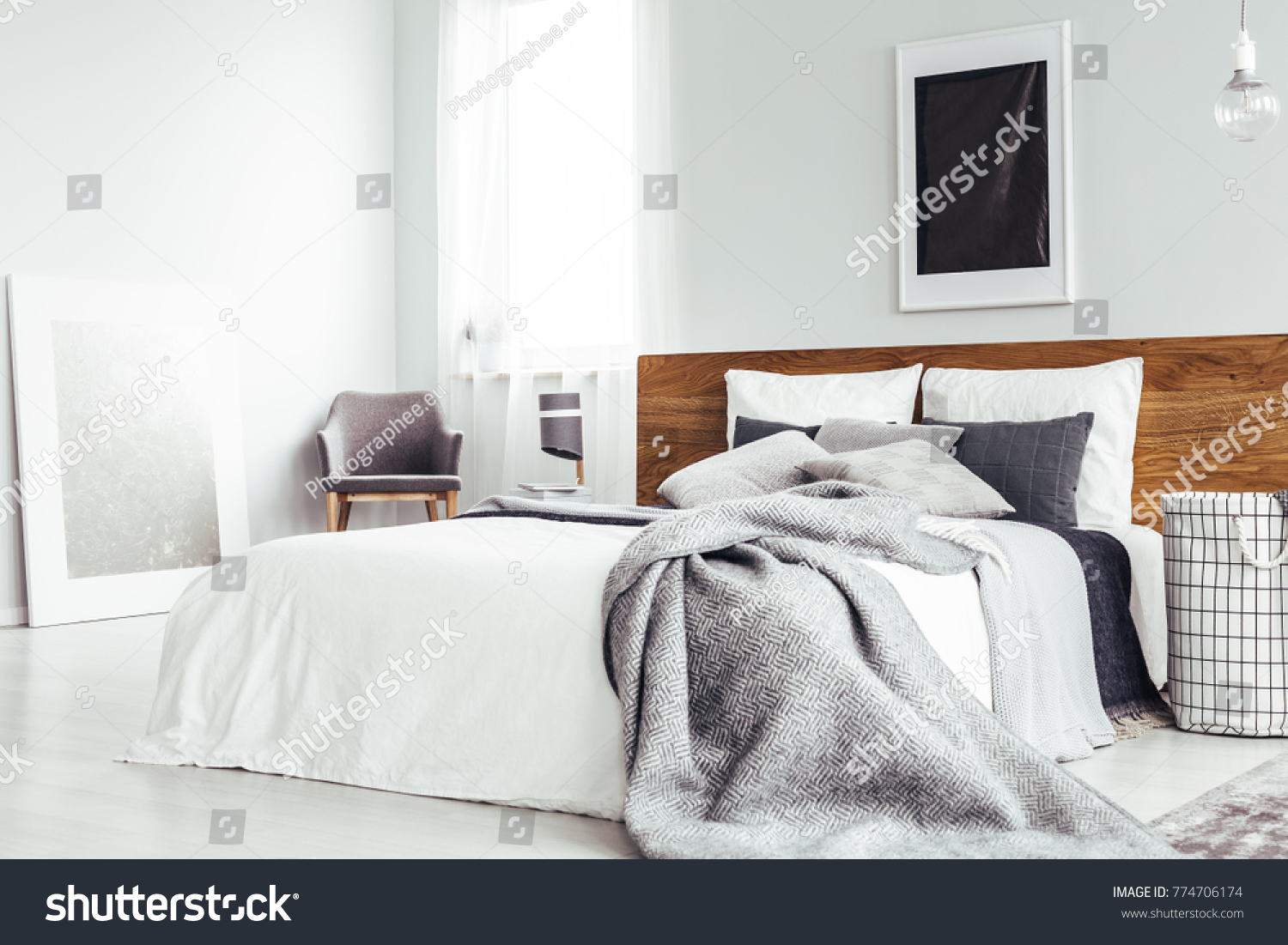 Grey blanket on bed with wooden bedhead in simple bedroom interior with dark poster and chair under window #774706174