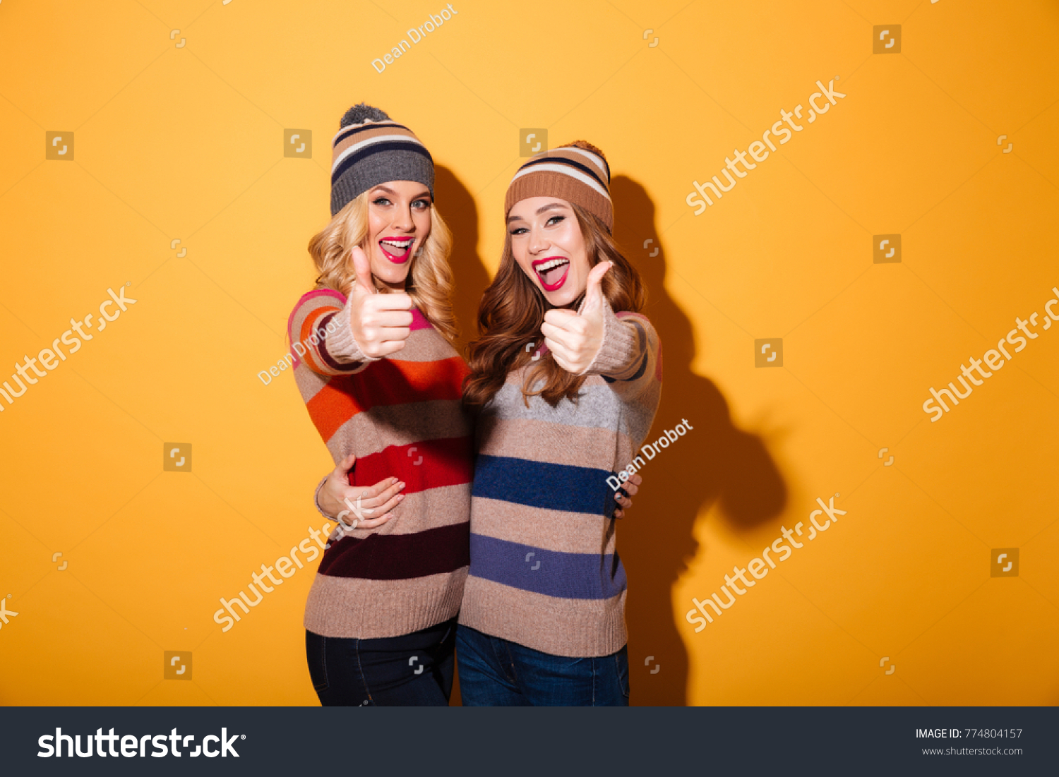 Portrait of two happy girls dressed in winter clothes standing and showing thumbs up gesture isolated over yellow background #774804157