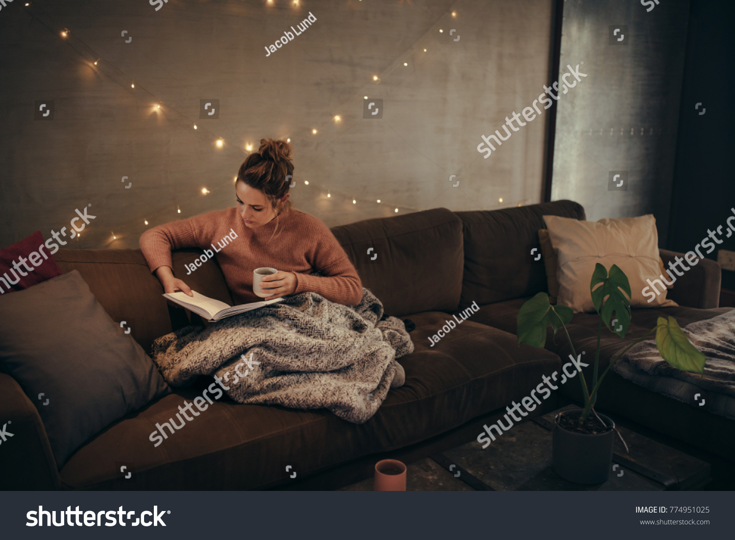 Young woman reading book and drinking coffee on sofa in hygge house. Caucasian female relaxing in cozy living room and reading a book. #774951025