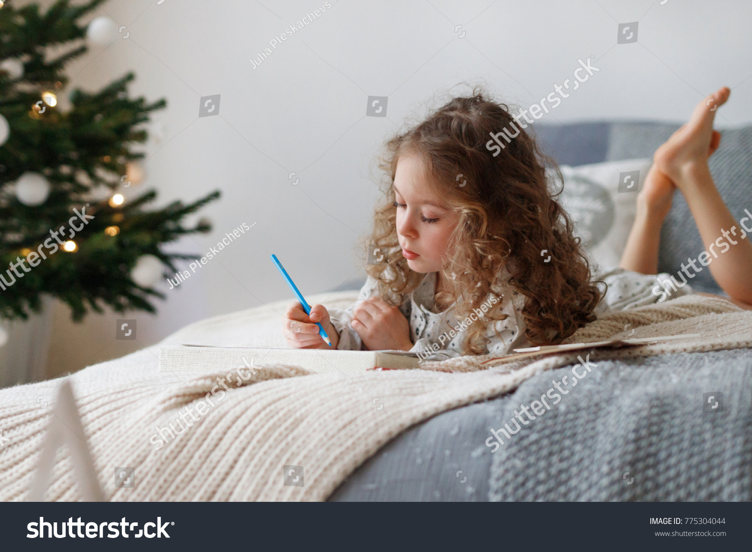 Indoor shot of attentive pretty small girl writes letter to Santa Claus before Christmas, thinks what present she expects, wants all dreams and wishes come true, lies on comfortable bed indoor. #775304044