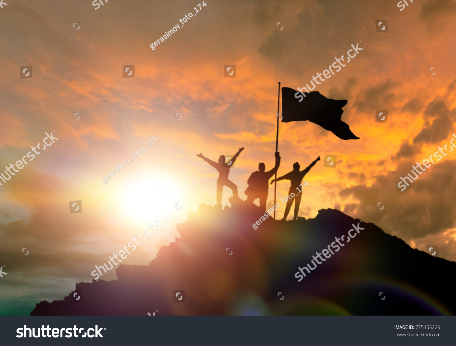 Conquest of height, silhouettes of three people, on top of a mountain, with a flag. Conceptual design. Against the background of the evening sky at sunset with clouds. #775455229