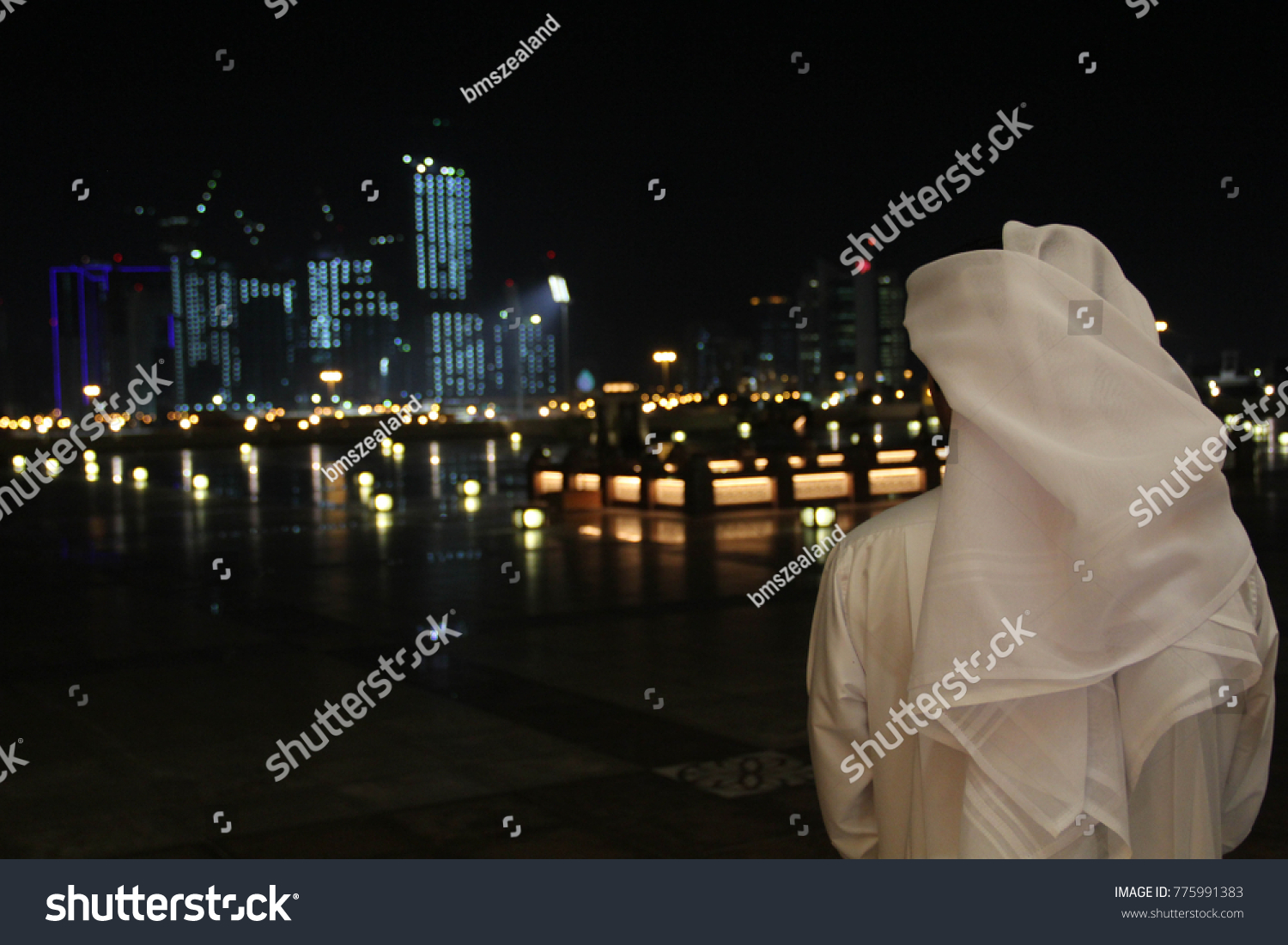 An Arab Qatari man wearing a white thobe or thwab (Arab garment) and white ghutra (or keffiyeh or headress) looking out to the evening or night skyline of Doha, Qatar #775991383