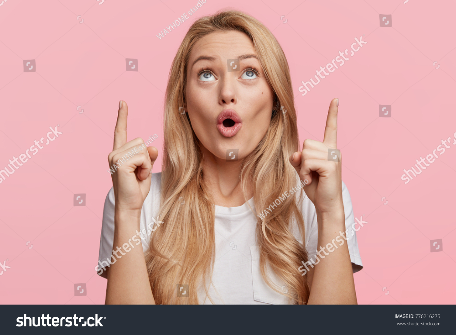 Good looking blonde female with pure healthy skin looks in amazement as indicates at something upwards, isolated over pink background. Pretty young woman sees amazing thing up, gestures indoor #776216275