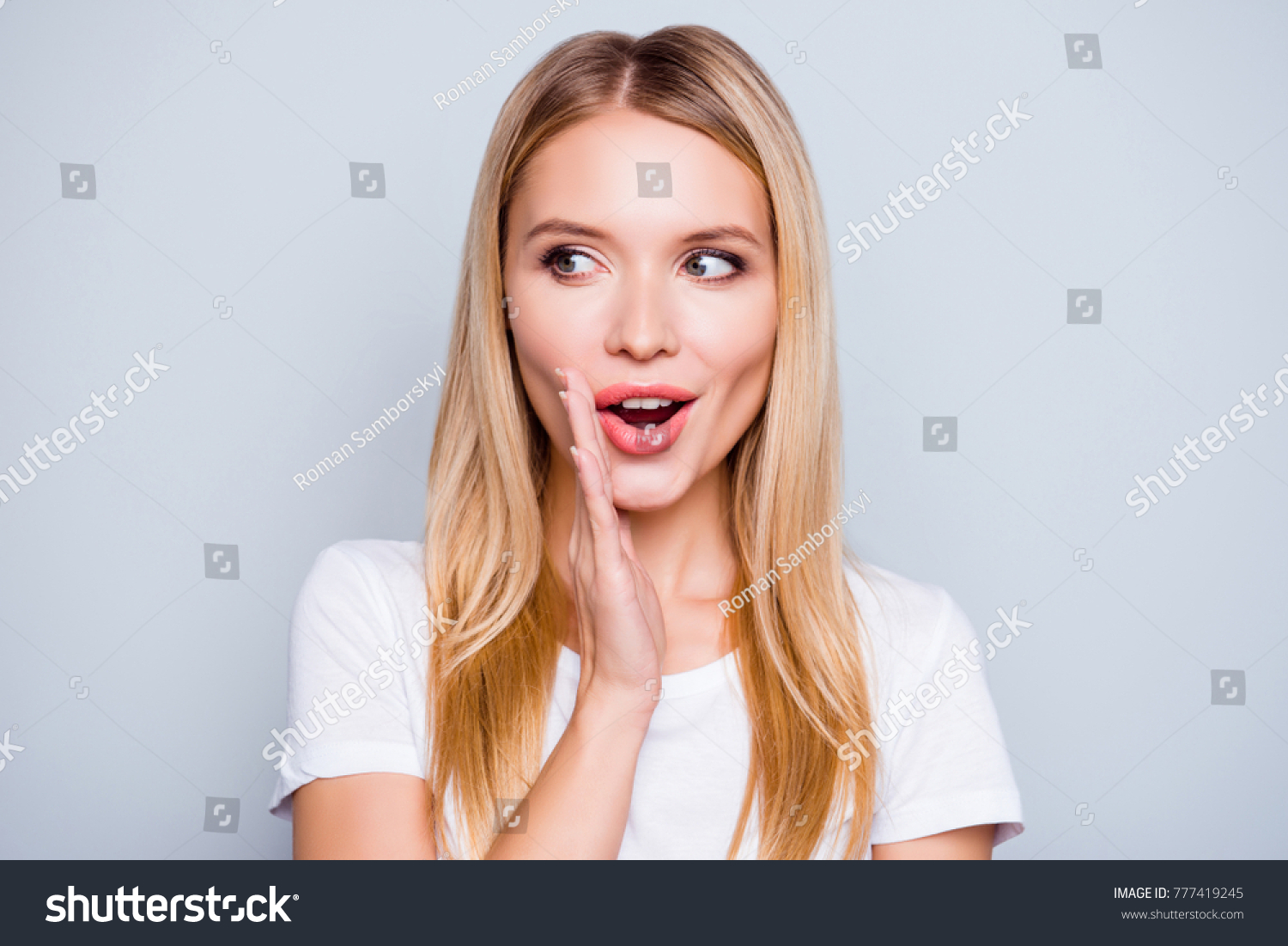 Don't tell anyone! Talkative mysterious pretty beautiful woman with blonde hair dressed in white tshirt is saying secret hot braking news and looking aside, isolated on grey background #777419245