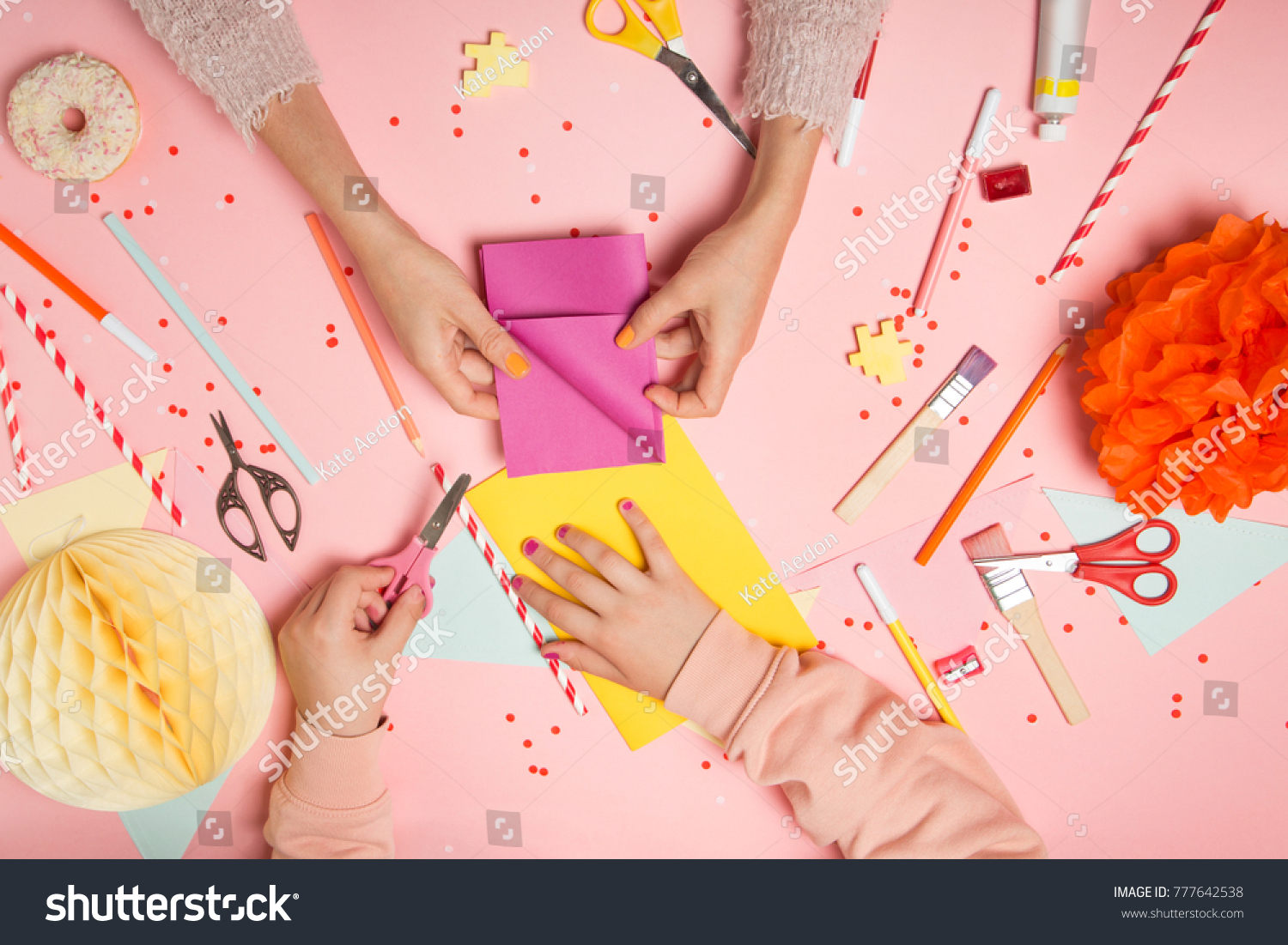 Colorful pink background with various party confetti, paper decoration, flags, stationary, DIY accessories with woman's and kid's hands making greeting card. Fat lay top view. Party arrangement #777642538