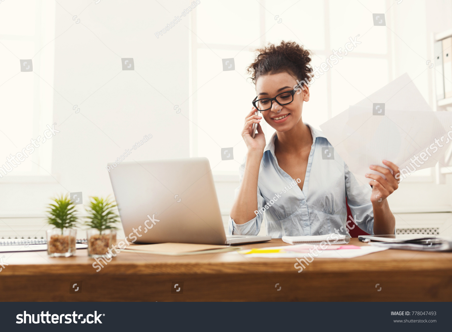 Happy african-american business woman talking on mobile at office. Young female manager consulting on phone, holding papers, using laptop at working place, copy space #778047493