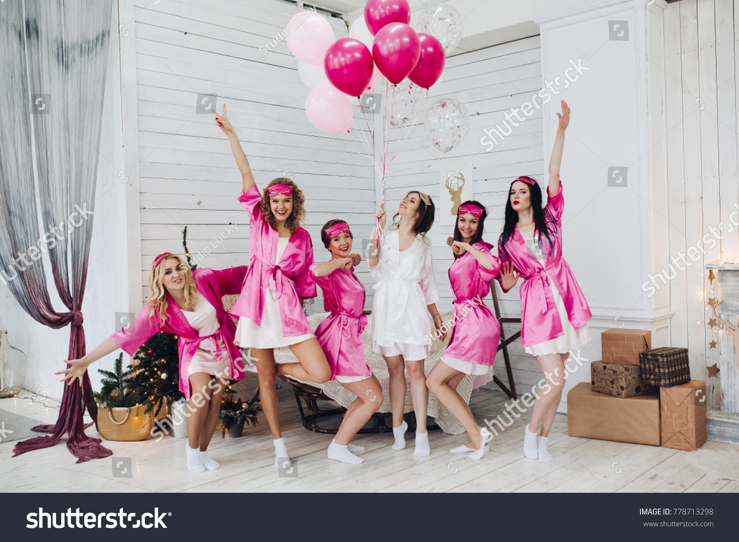Full length portrait of happy lovely girlfriends in pink robes and sleeping masks jumping and having fun at hen party. Bride-to-be in white robe holding pink air balloons with smile standing on tiptoe #778713298
