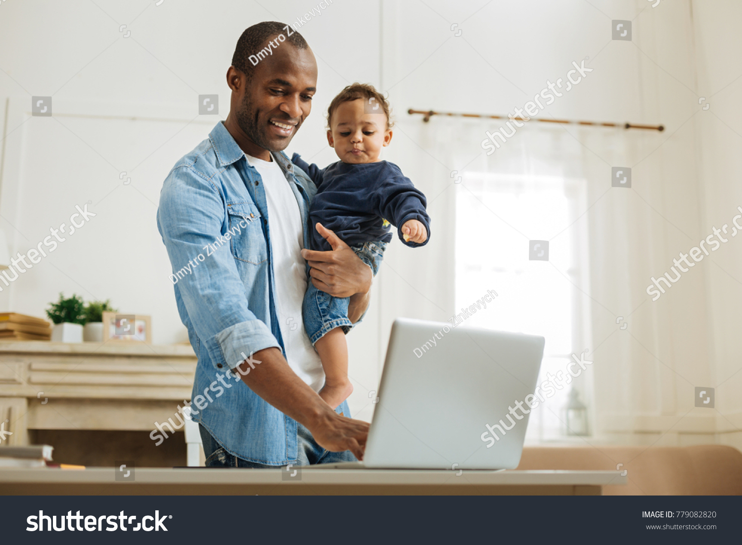 Typing. Attractive alert young afro-american father holding his little son and typing on the laptop while standing at the table and a fireplace in the background #779082820