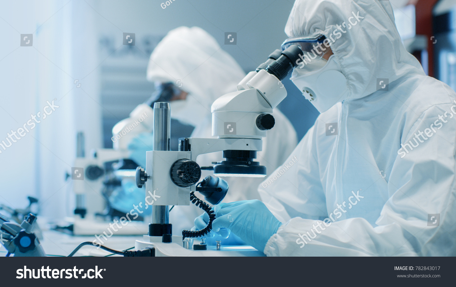 Two Engineers/ Scientists/ Technicians in Sterile Cleanroom Suits Use Microscopes for Component Adjustment and Research. They Work in an Electronic Components Manufacturing Factory. #782843017