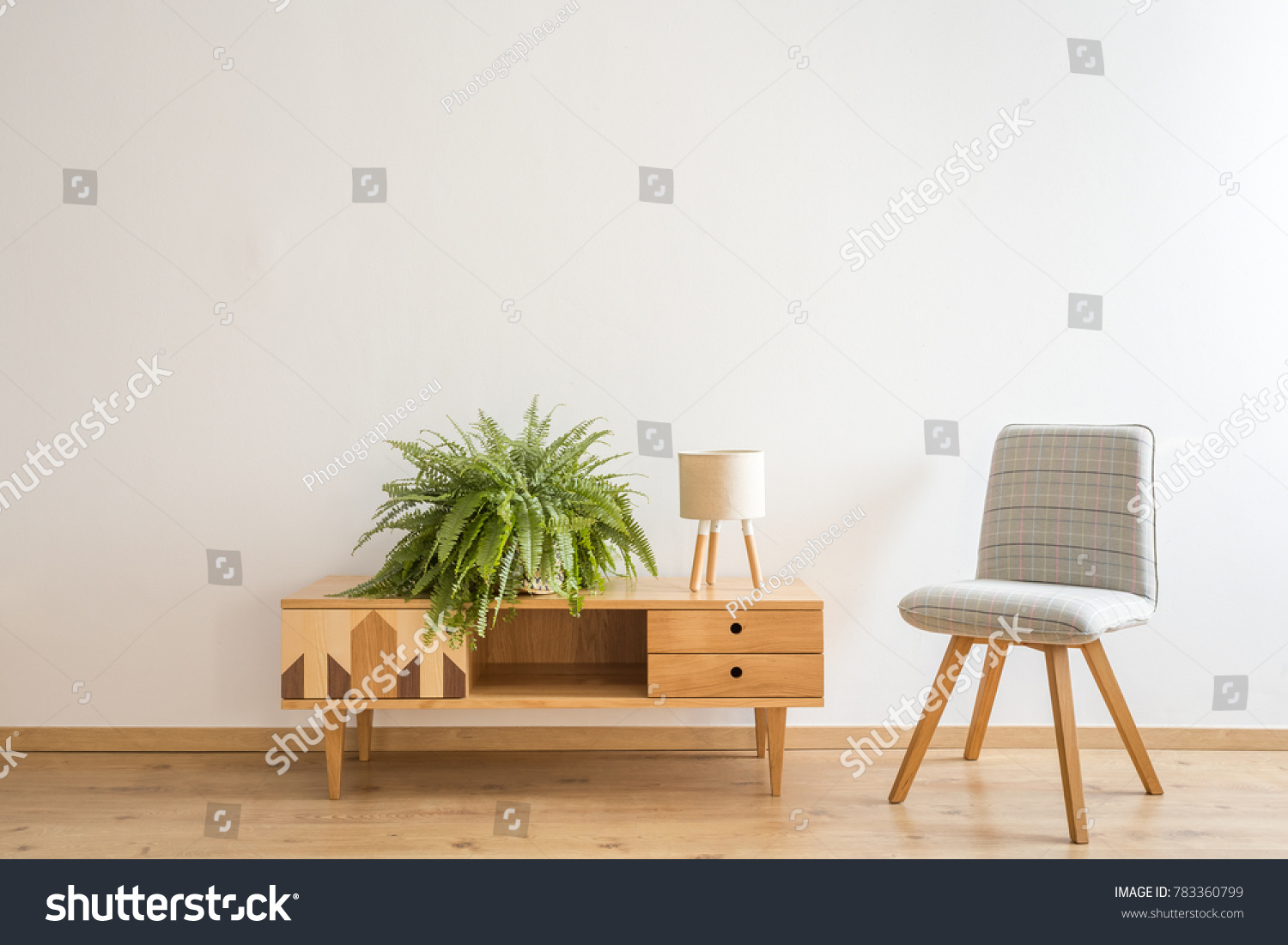 Simple, gray chair stranding next to a wooden cupboard with small lamp and fern on it #783360799