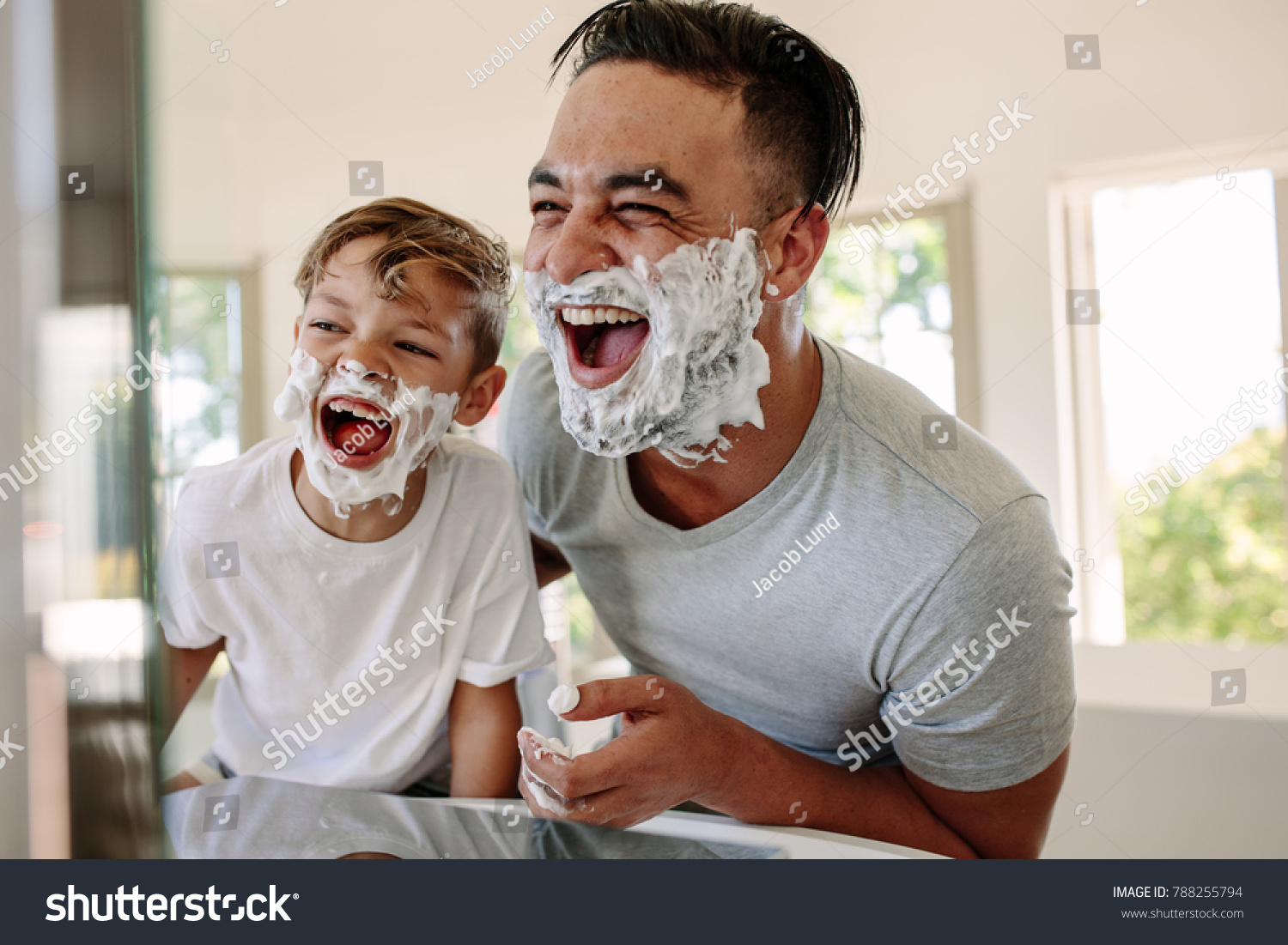 Man and little boy with shaving foam on their faces looking into the bathroom mirror and laughing. Father and son having fun while shaving in bathroom. #788255794