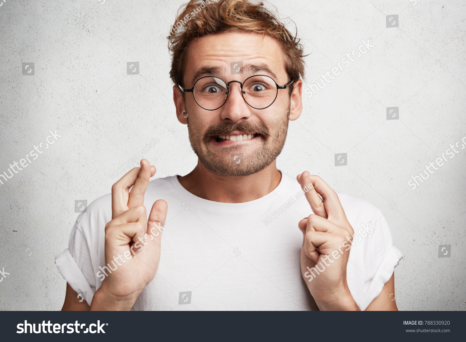 Positive bearded young male wears round spectacles, crosses fingers and bites lips has great hope for better, wishes something, isolated over white concrete wall. People, wish and desire concept #788330920