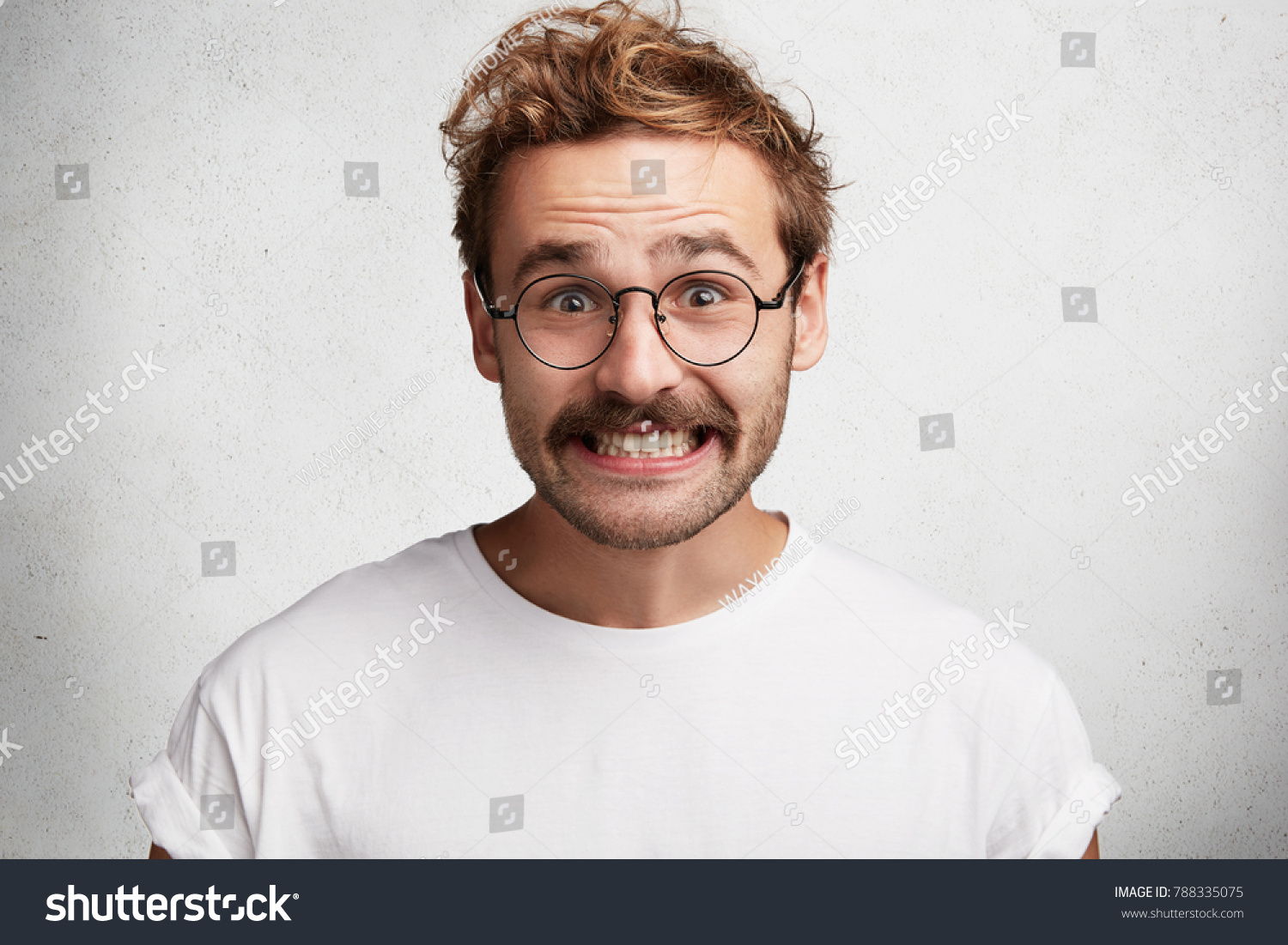 Funny attractive bearded young male with comic smile, has awkward look, pretends to be happy, looks gladfully through round spectacles, laughs at joke, isolated over white concrete background. #788335075