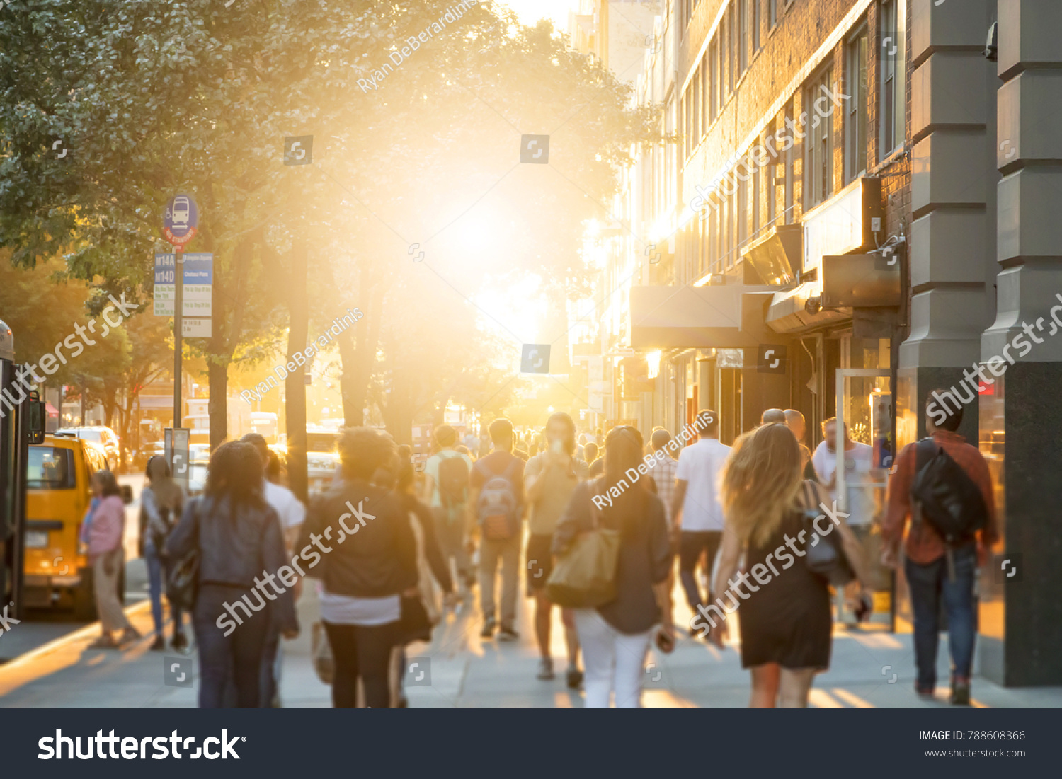 Man stands in the middle of a busy sidewalk looking at his cell phone while crowds of people walk around on 14th Street in Manhattan, New York City with the glow of sunlight in the background. #788608366