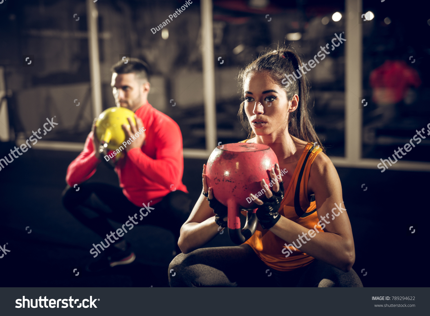 Close up of focused and motivated strong young fitness couple in sportswear crouching with the kettlebells reversed and doing squats in the gym. #789294622