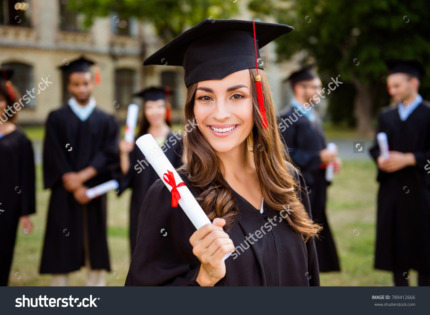 Happy cute brunette caucasian grad girl is smiling, blurred class mates are behind. She is in a black mortar board, with red tassel, in gown, with nice brown curly hair, diploma in hand #789412666
