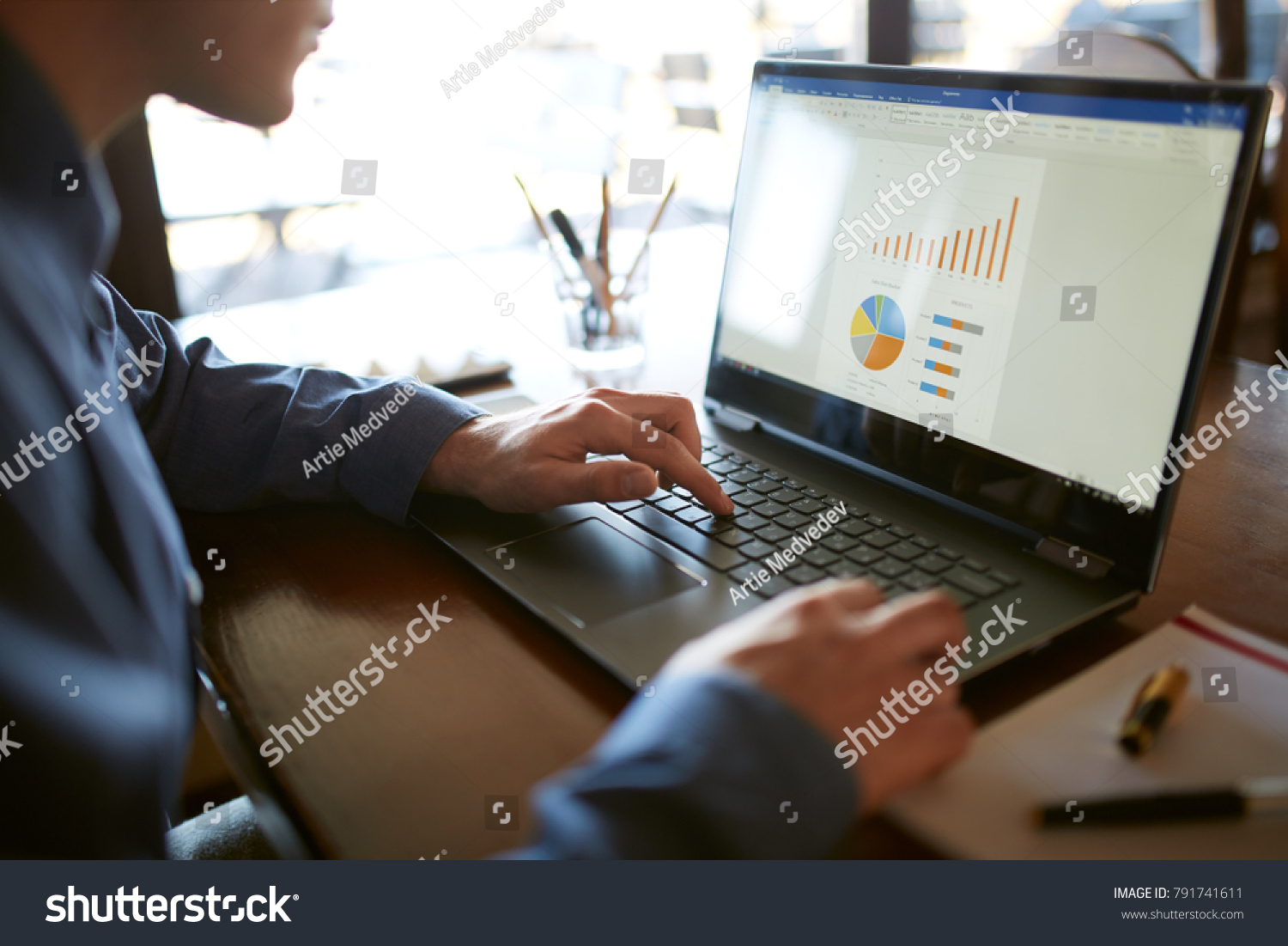 Close-up back view of caucasian businessman hands typing on laptop keyboard and using touchpad. Notebook and pen on foreground of workspace. Charts and diagrams on screen. Isolated no face view. #791741611