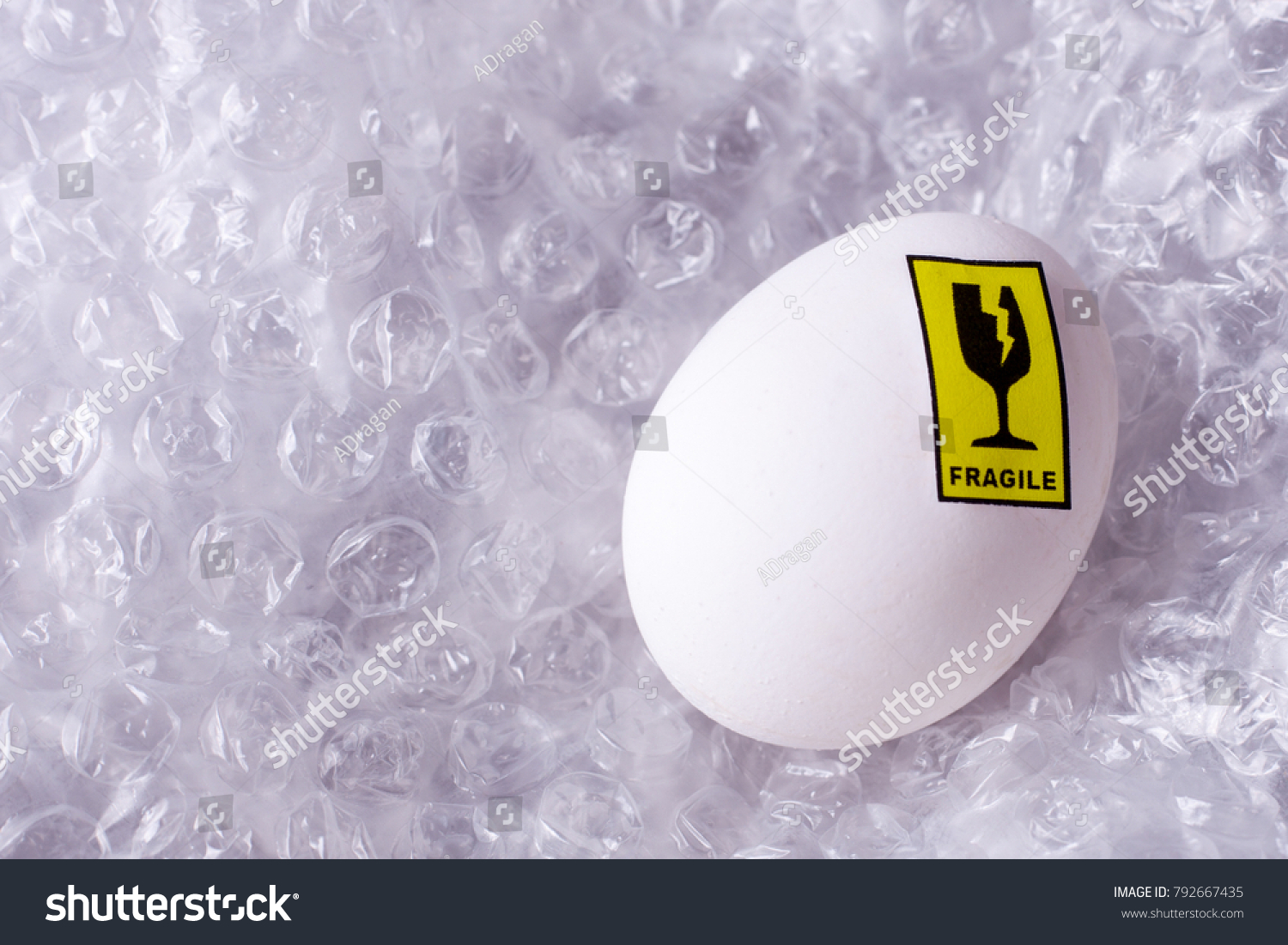 Delivery of fragile goods in the package. Egg with a sticker with a symbol of a glass with a crack and an inscription fragile in a film with air pads. #792667435