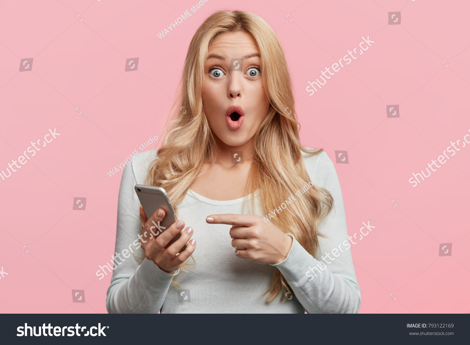 Blonde young female with pleasant appearance looks with terrified expression in smart phone, reads shocking news on webpage, isolated over pink background. Woman indicates at digital telephone #793122169