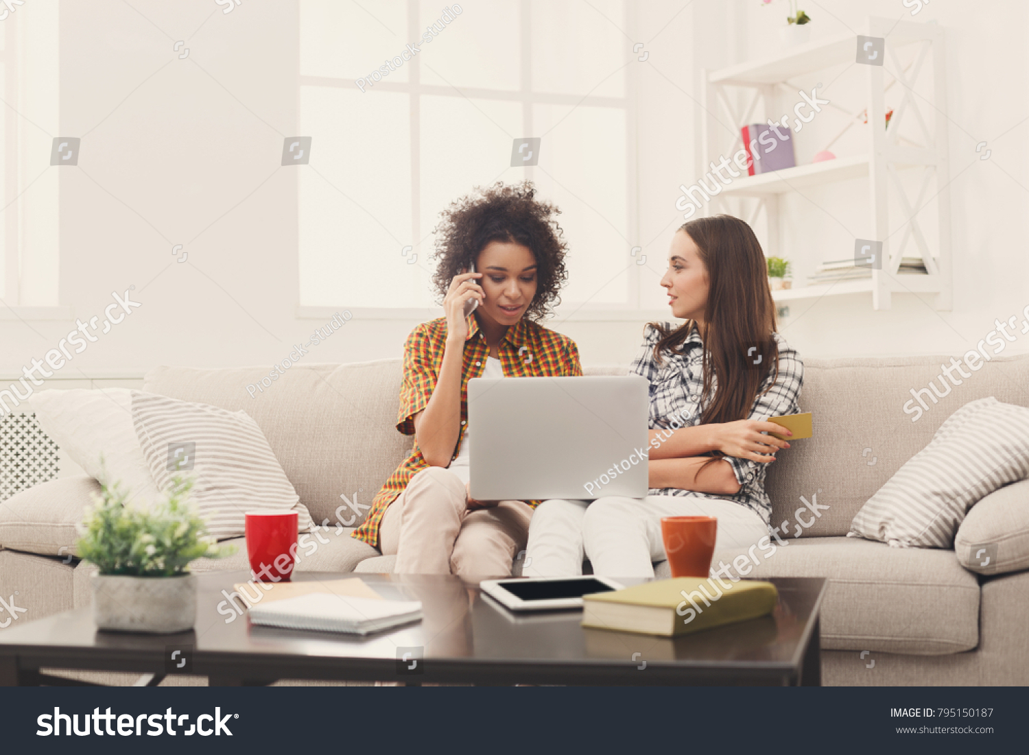 Two happy women shopping online with credit card and laptop sitting on sofa at home and making order by mobile phone, copy space #795150187