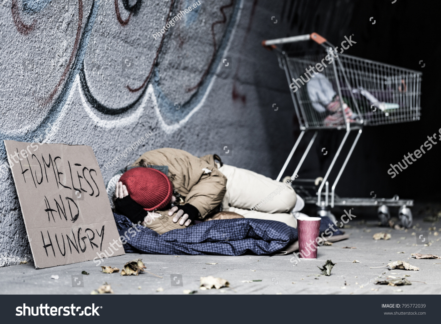 Dirty tramp lying on blanket next to a sign and trolley in the shelter of the city #795772039