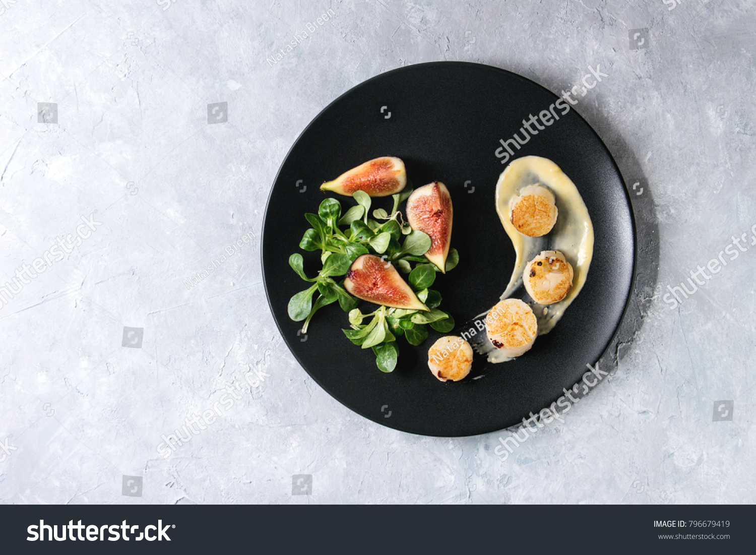 Fried scallops with lemon, figs, sauce and green salad served on black plate over gray texture background. Top view, copy space. Plating, fine dining #796679419