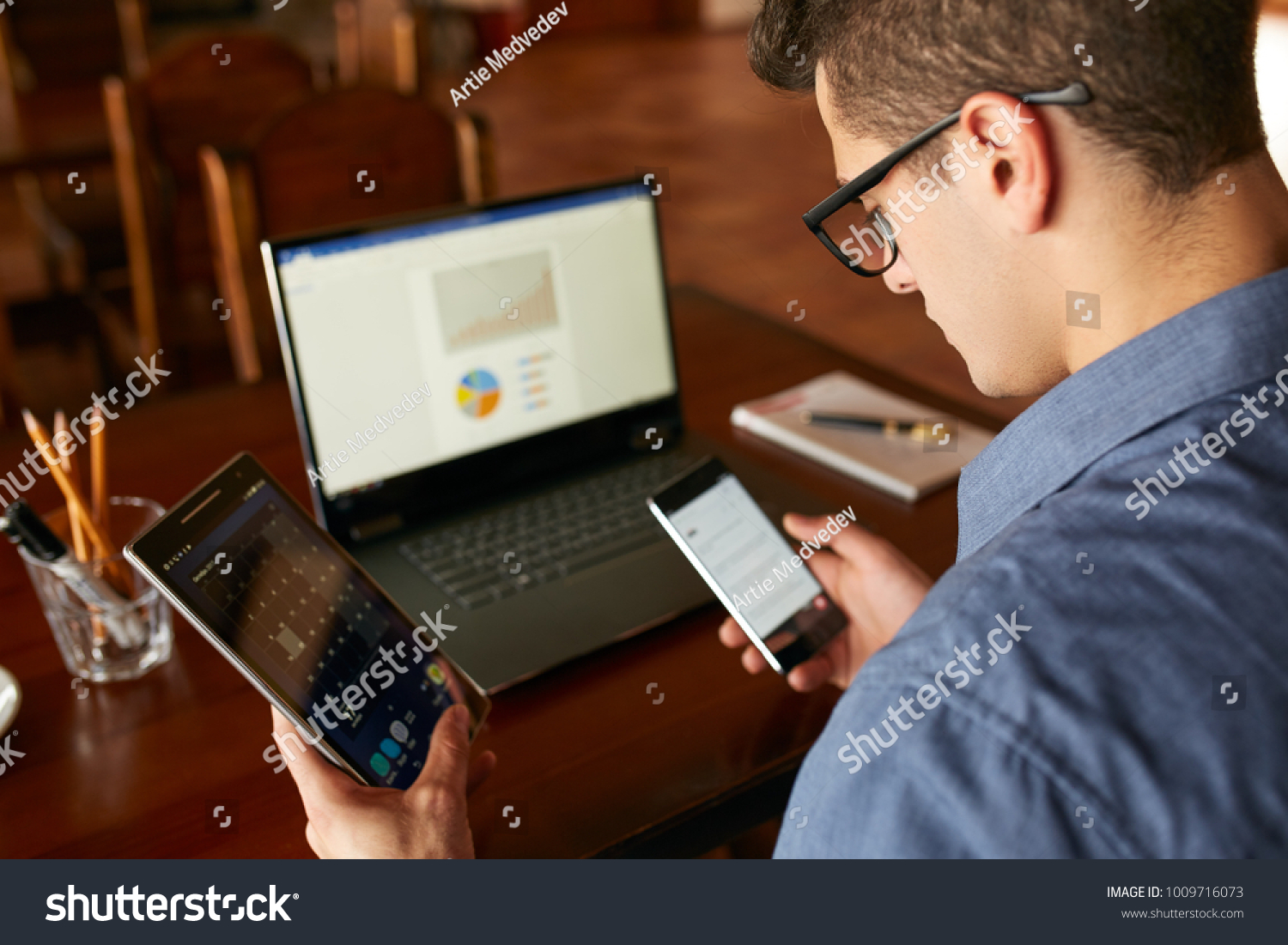 Attractive man in glasses working with multiple electronic internet devices. Freelancer businessman has laptop and smartphone in hands and laptop on table with charts on screen. Multitasking theme. #1009716073