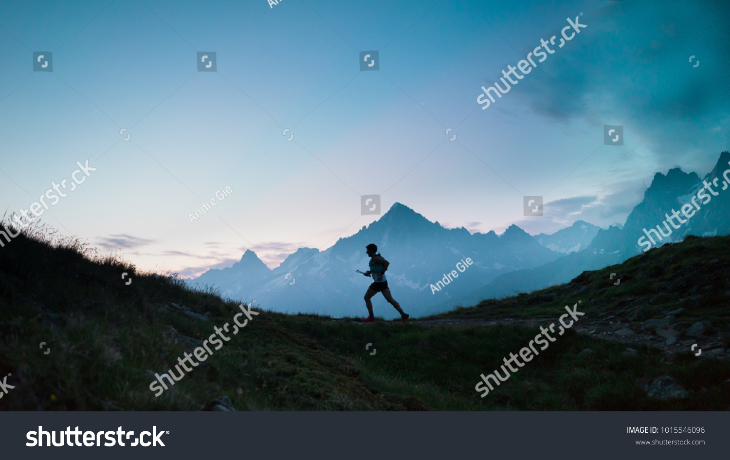 A single trail runner, silhouetted against the sky at sunrise while running in the mountains of the Alps along a steep trail #1015546096