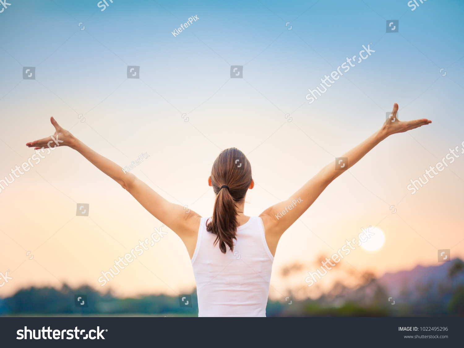Young woman raising her arms up against the sunset feeling free.  Happiness and joy concept.  #1022495296