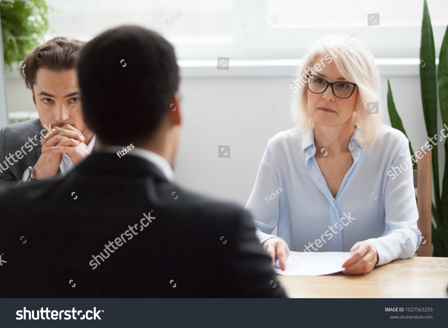 Serious attentive senior female hr manager employer listening to candidate at job interview, focused strict mature businesswoman thinking about hiring decision at difficult group negotiations concept #1027563259