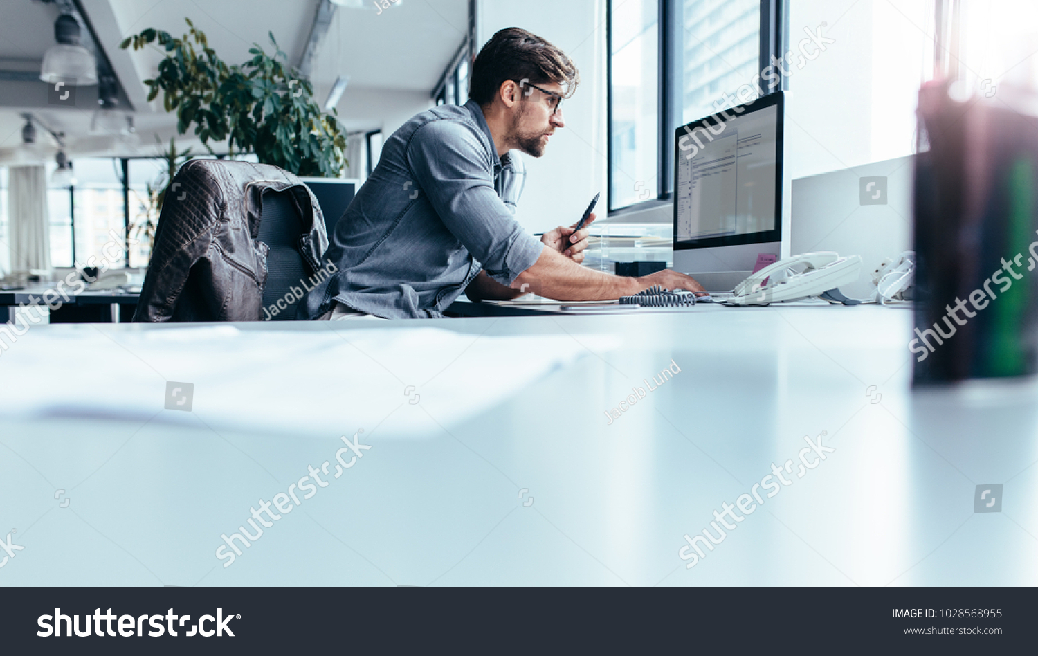 Young man sitting in office and working on desktop pc. Businessman looking at computer monitor while working in office. #1028568955
