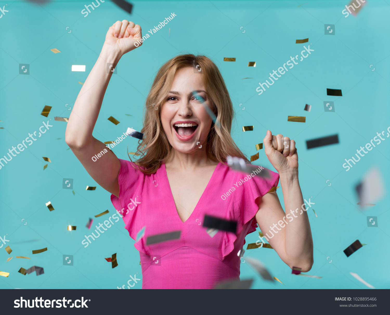 Portrait of contented blonde girl exulting while raising her hands up as gesture of triumph. Confetti flying in air. Isolated on blue background #1028895466