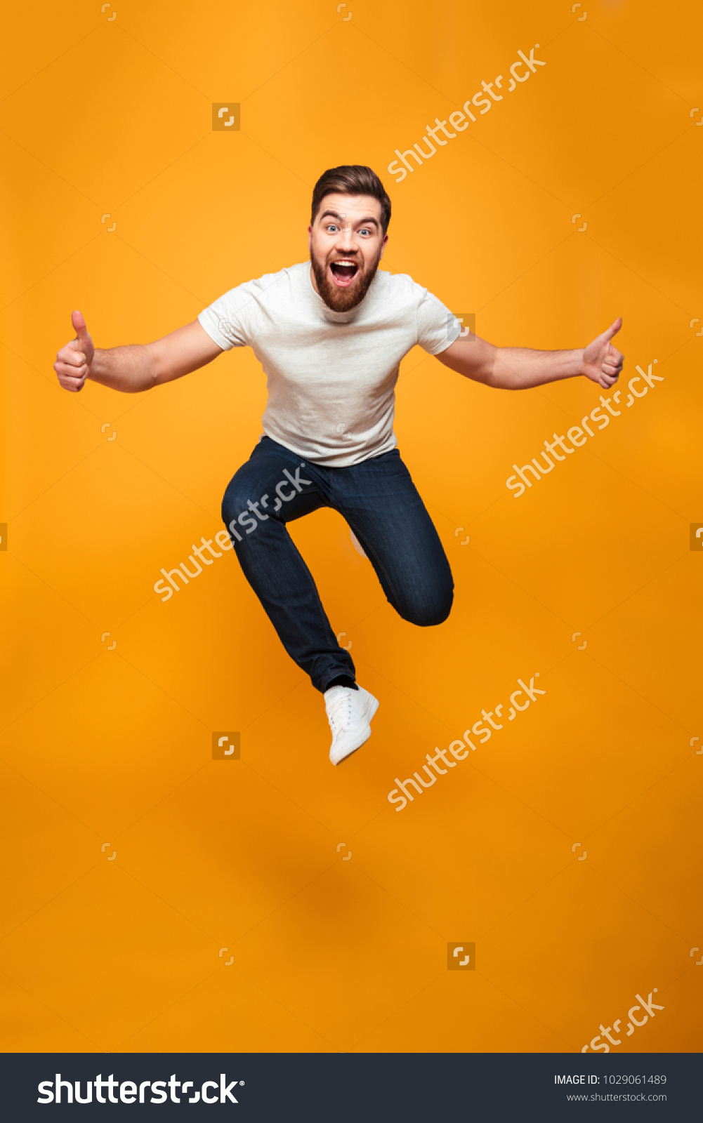 Full length portrait of an excited bearded man jumping and showing thumbs up isolated over yellow background #1029061489