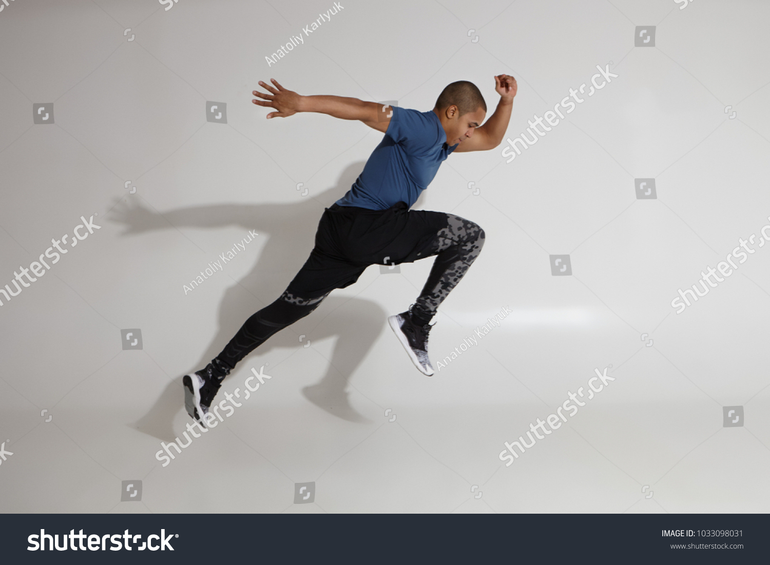 Endurance and stamina. Side view of athletic young Afro American sportsman training in studio, doing high jumps. Action shot of serious determined dark skinned male in sportswear jumping at white wall #1033098031